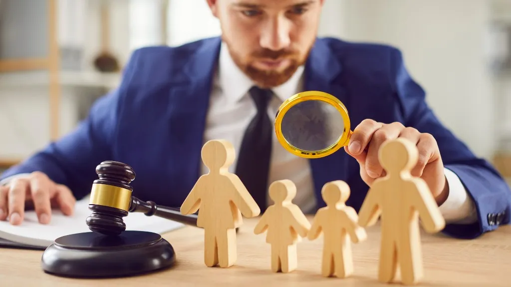Man in blue suit examining wooden family figures with magnifying glass, gavel on desk.
