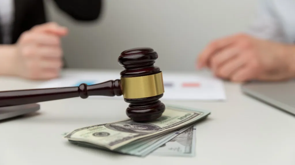 Wooden judge gavel resting on a stack of US dollar bills with blurred hands in background.