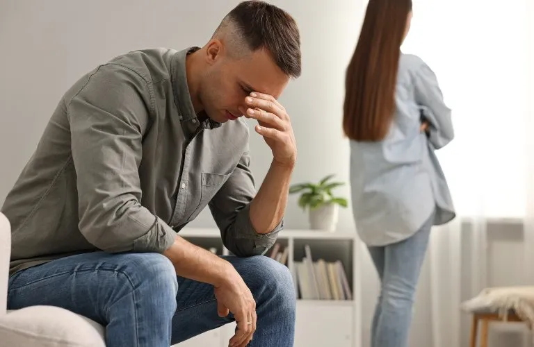 Man in gray shirt and jeans sitting with head in hand, woman standing with back turned.
