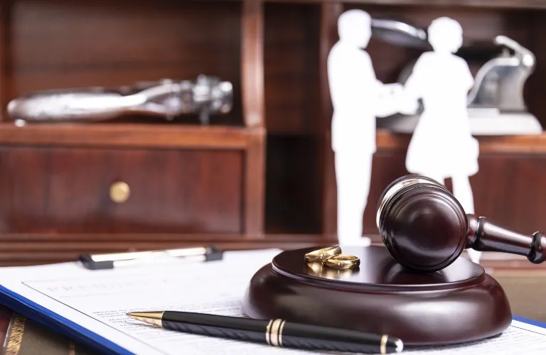 Wooden judge's gavel with two gold rings, pen, and blurred cutout figures of a couple on desk.
