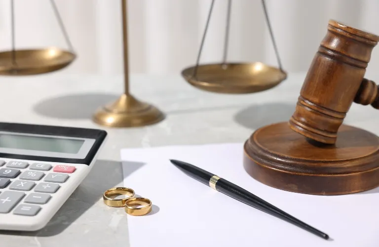 Wooden judge's gavel, brass scales of justice, two gold rings, calculator, pen, and blank paper.