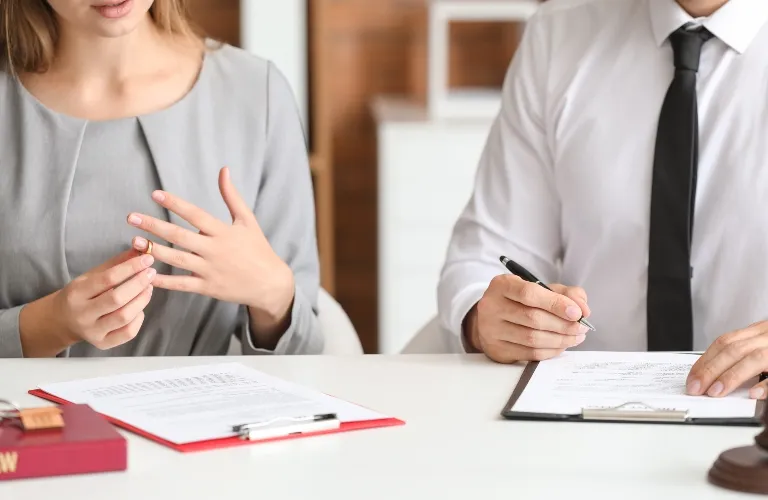Two professionals at desk, woman removing ring, man writing on clipboard, legal documents present.