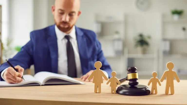 Man in blue suit writing in a book with wooden family figures and a judge's gavel on the table.