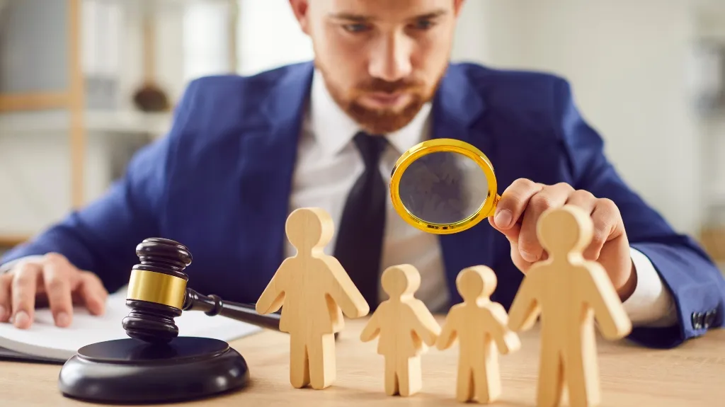 Man in blue suit examining wooden family figures with magnifying glass, gavel on desk.