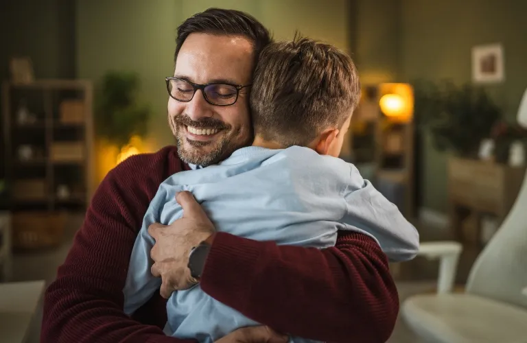 Man wearing glasses and a maroon sweater embraces a young boy in a light blue shirt indoors.