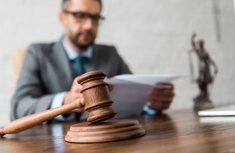 Wooden judge's gavel on desk with blurred man in suit reading documents in background.
