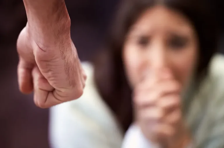 Close-up of a clenched fist in the foreground with a blurred woman appearing distressed in the background.