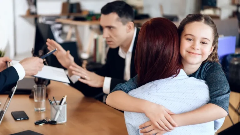 Young girl smiling while hugging a woman in an office setting with two men discussing documents.