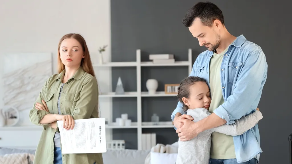 Woman holding a divorce decree with crossed arms, a man hugging a child in a living room.