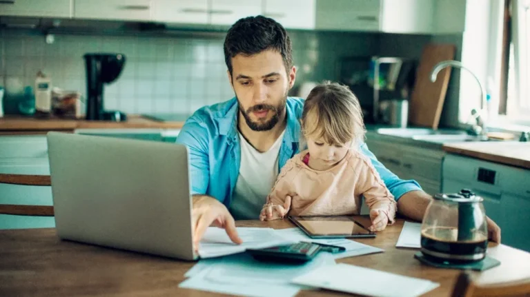 Man in blue shirt working on laptop with young child using tablet in kitchen.