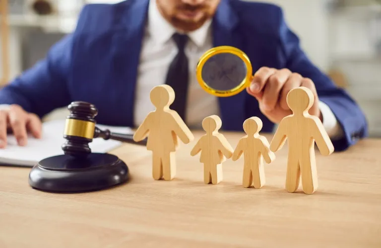 Man in blue suit examining wooden family figures with magnifying glass beside judge's gavel.