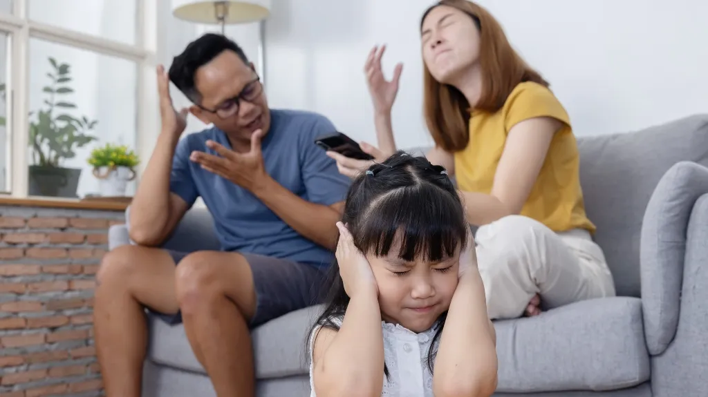 Young girl covering ears in distress with parents arguing in the background on sofa.