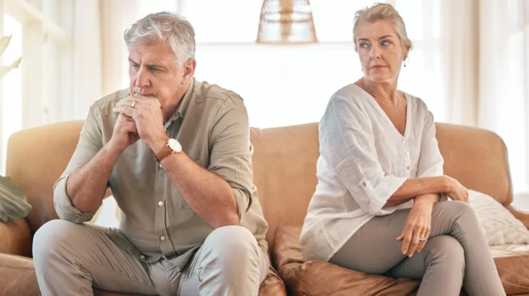 Mature man and woman sitting apart on a beige couch, appearing upset and distant.