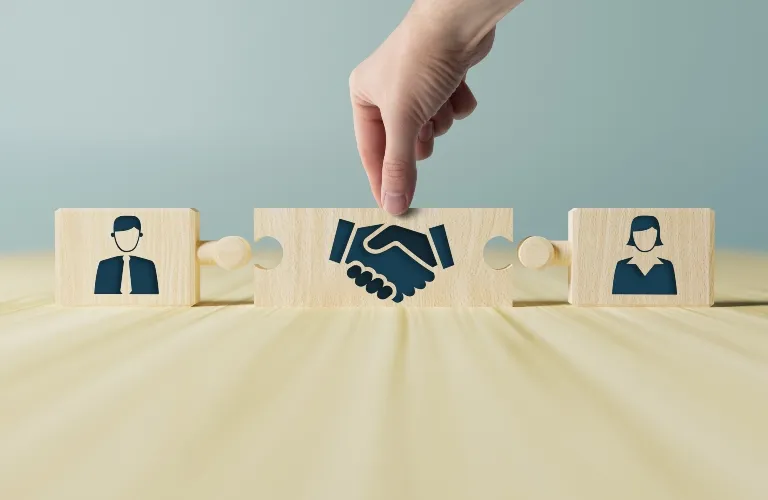 Hand placing wooden puzzle piece with handshake icon between male and female businessperson icons.
