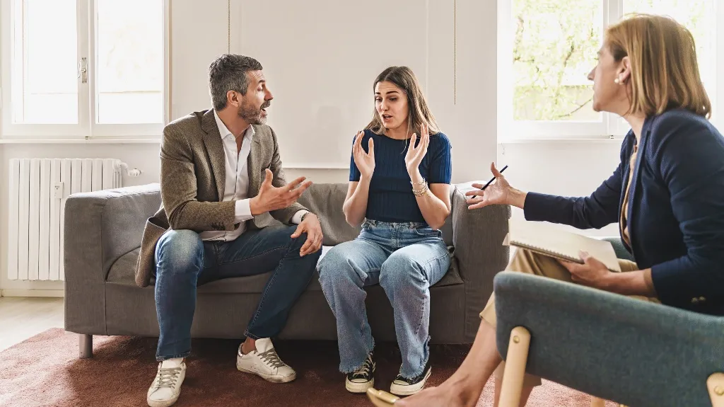 Three adults engaged in a serious conversation in a bright room, seated on gray sofas.