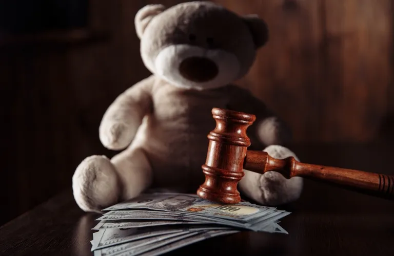 Wooden judge's gavel resting on a stack of US dollar bills with a teddy bear in the background.