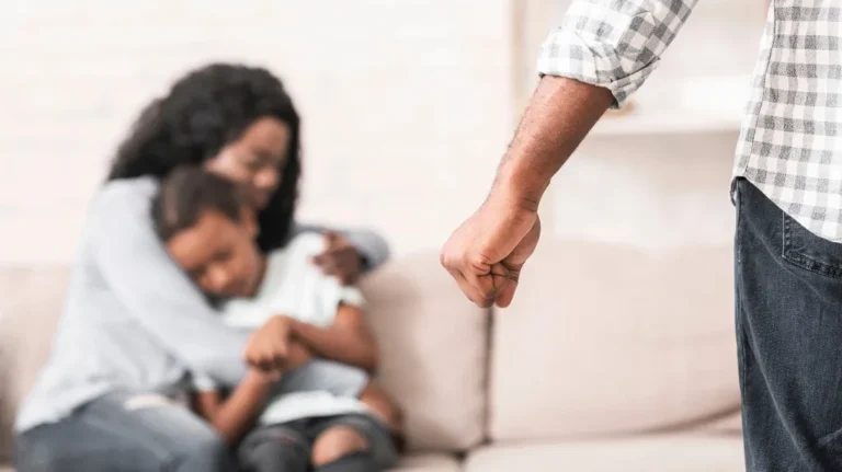 Close-up of a clenched fist in foreground with a woman comforting a child on a sofa in the background.