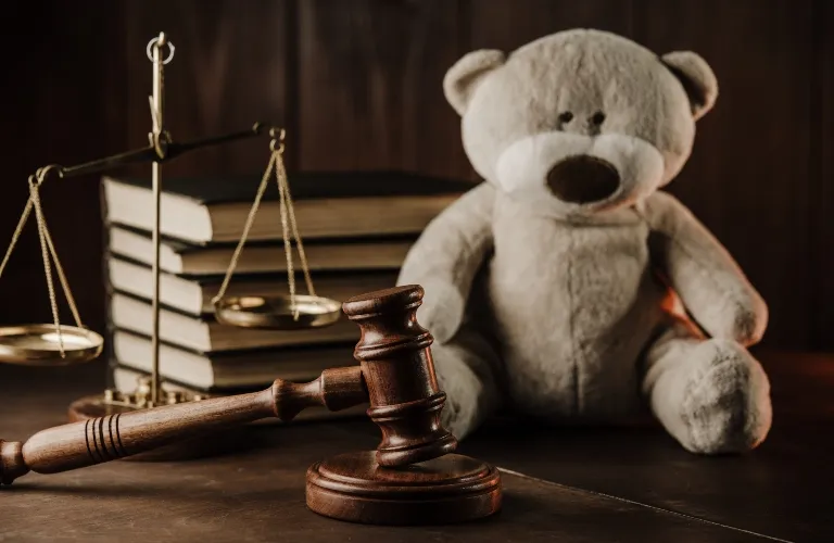 Wooden judge's gavel, brass scales of justice, stacked books, and a beige teddy bear on a wooden surface.
