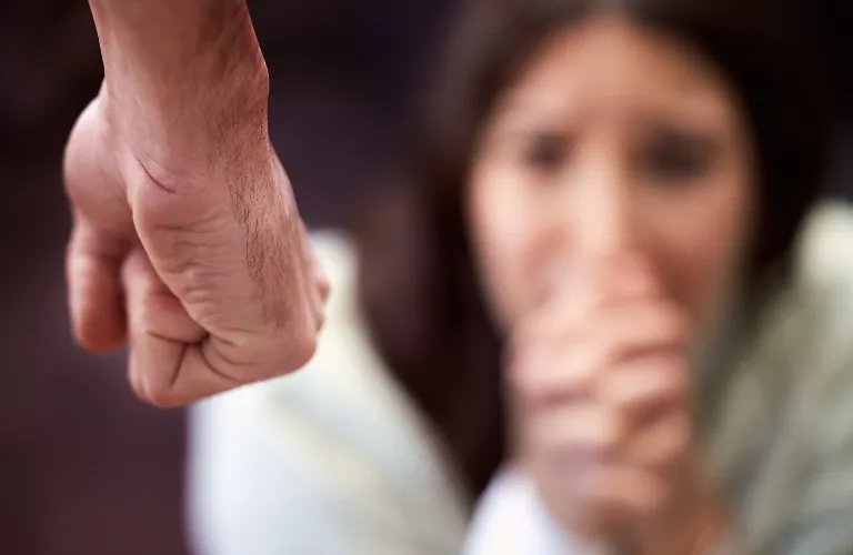 Close-up of a clenched male fist in the foreground with a blurred woman appearing fearful in the background.