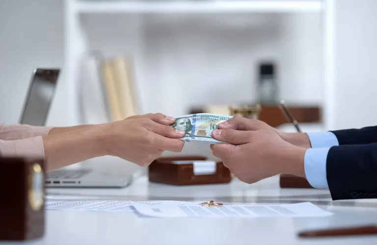 Two people exchanging a stack of US hundred-dollar bills over a white office desk with documents and a laptop.