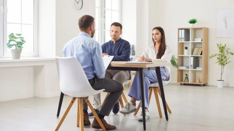 Three professionals engaged in a discussion around a wooden table in a bright office.