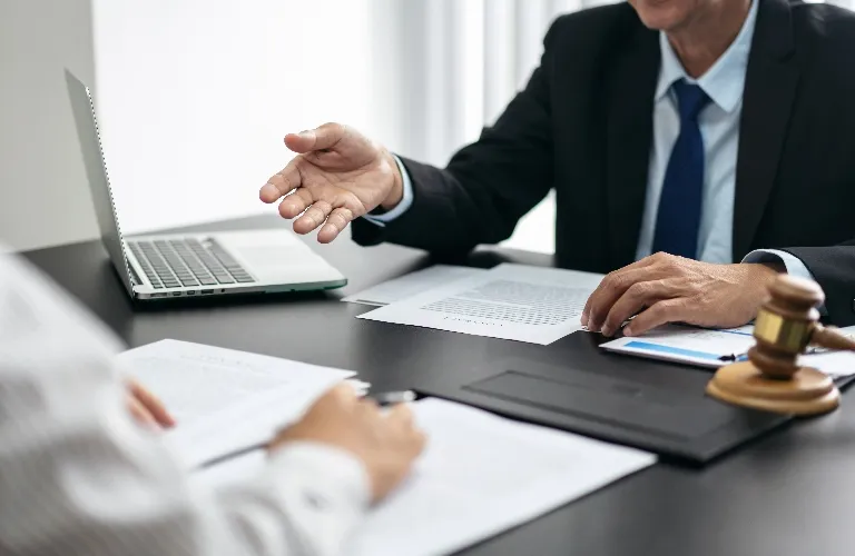 Two professionals in formal attire discussing documents at a desk with a laptop and a wooden gavel.