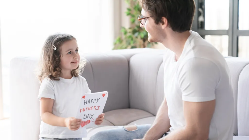 Young girl presenting handmade Father's Day card to smiling man in white shirt on sofa.