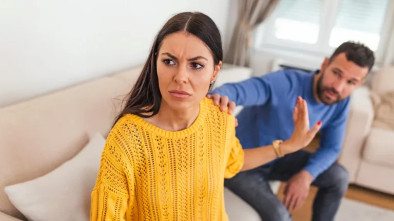 Woman in yellow sweater rejecting man in blue shirt with raised hand on beige couch.