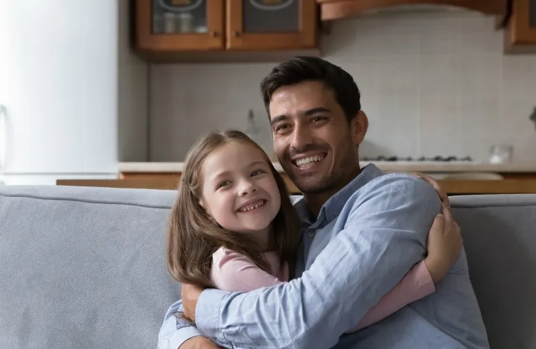 Man in a blue shirt and young girl in a pink shirt smiling and hugging on a gray couch.