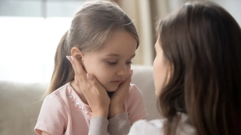 Woman gently holding young girl's face, comforting her in a softly lit room.